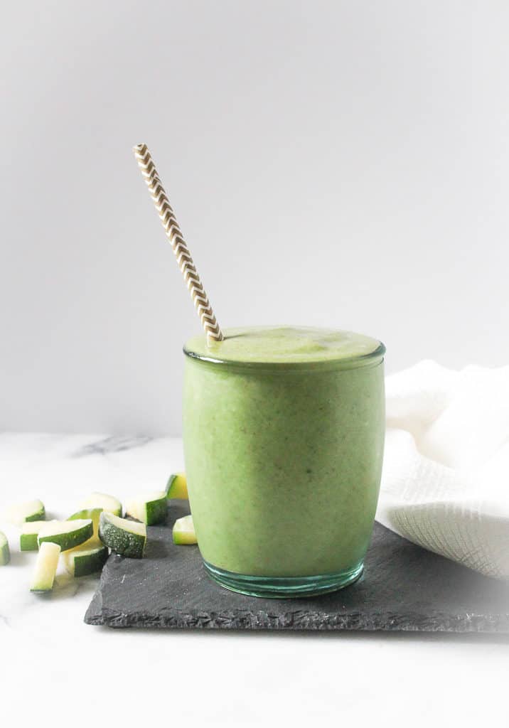healthy zucchini smoothie in a small glass with paper straw next to diced zucchini pieces