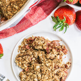 strawberry rhubarb crisp on small white plate next to baking dish and whole strawberries