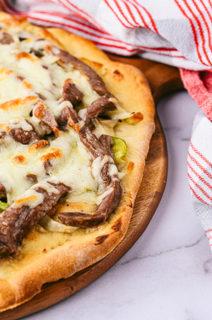 homemade philly cheesesteak pizza next to a red and white dish towel