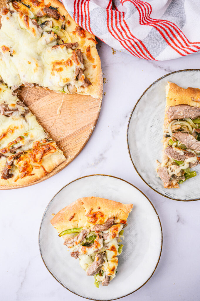 top view of philly cheesesteak pizza on two plates next to the whole pizza